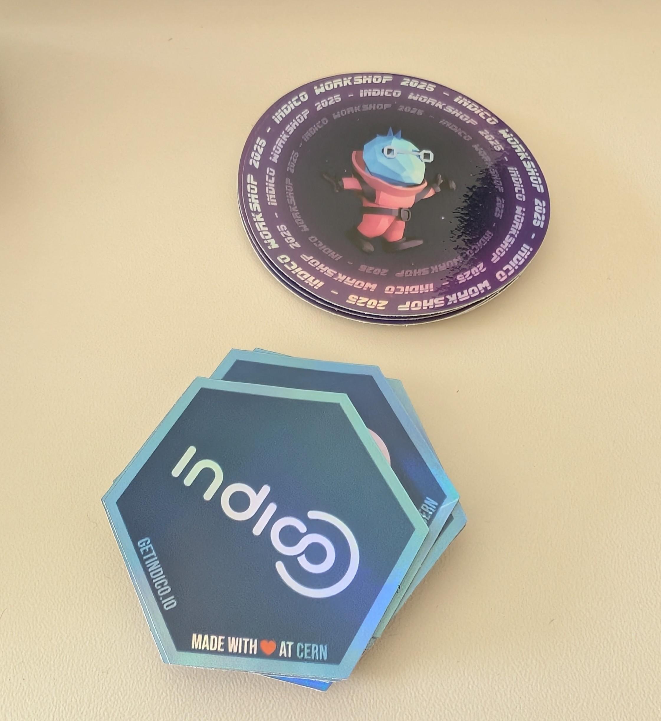 Hex and round Indico stickers: the former has an indico logo on blue background, the other one a low poly version of the Indico dinosaur in an astronaut suit, floating in the void, the purple border says "Indico Workshop 2025". They're both foil finished.